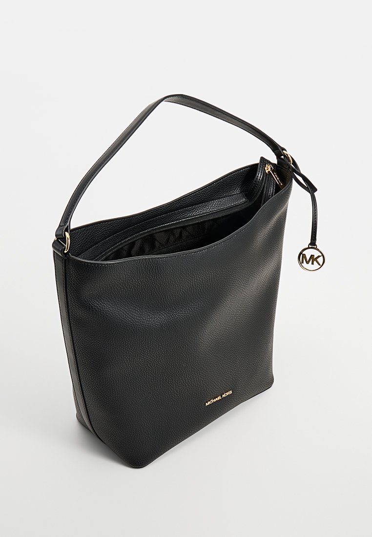 Borsa a spalla in pelle nera con texture, zip dorata, logo Michael Kors e charm rotondo MK su una tracolla, su uno sfondo grigio chiaro uniforme.