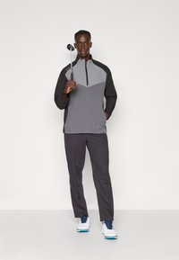Golfbekledd set med halv-zip, todelt grå genser og mørke grå bukser, kombinert med hvite og blå golf sko, holder en golfklubb.