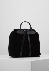 Černá semišová kabelka typu bucket s koženou rukojetí a popruhy, vystavena na bílém podstavci na jednolité šedé pozadí.