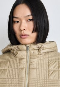 Beige quilted jacket featuring a hood, front zipper, and drawstring accents. Textured pattern throughout with a smooth finish.