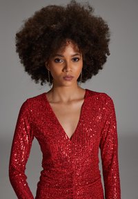 Rode pailletten jurk met een diepe V-hals en lange mouwen, kenmerken een getailleerde silhouet en een textuurpatroon van glanzende pailletten.