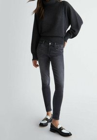 Suéter gris de cuello alto con puños y dobladillos acanalados, combinado con jeans oscuros ajustados y mocasines negros con acentos blancos y herrajes dorados.