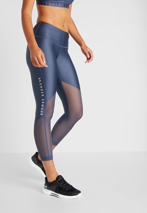 Vrouw draagt marineblauwe Under Armour leggings met meshpanelen en zwarte sportschoenen tegen een effen lichte achtergrond.