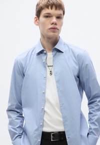 Hele sinine nööbitav särk klassikalise kraega, kantud valge T-särgi peal, sobitatud musta vööga. Sile kangas, nähtavaid mustreid ei ole.