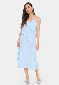 Rochie midi de un albastru deschis, cu accente volănoase pe o parte, bretele subțiri și o textură moale. Purtați cu pantofi cu accent de fundiță argintie.