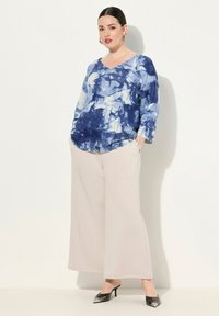 Top bleu tie-dye à col en V avec des manches trois-quarts, associé à un pantalon large beige et des escarpins noirs à bout pointu. Textures lisses.