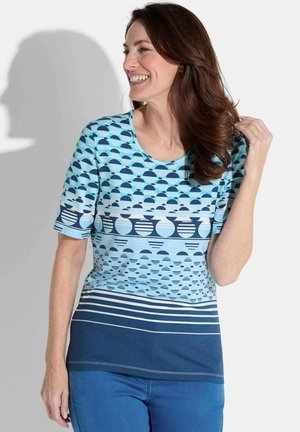 Femme souriante, portant une chemise bleue à manches courtes avec des motifs géométriques de cercles et de rayures, et un jean bleu, touchant ses cheveux.