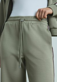 Ljus olivgröna sweatpants med elastisk midja, dragkedja, sidofickor och murgröna sidostrimmor. Slät, texturerad tyg.