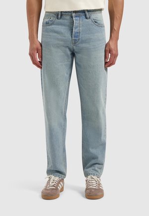 Lichtblauwe denim jeans met een relaxte pasvorm, voorzien van vijf zakken en een ritsluiting. Gecombineerd met bruin suède schoenen en witte veters.