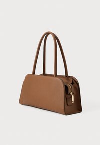 Bolsa de couro castanho com duas alças longas e detalhe em zíper dourado, de pé sobre um fundo claro e simples.