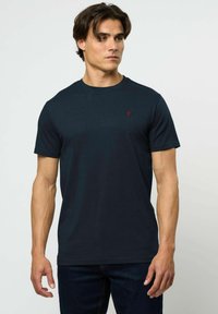 Jauns vīrietis ar īsiem tumšiem matiem, tērpies vienkāršā tumši zila T-kreklā un tumšās džinsās, stāvot pret baltu fonu, skatoties pa labi.