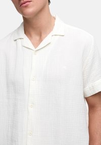 Camisa blanca de manga corta en un material texturizado y ligero. Presenta un cierre frontal con botones y un logo sutil en el lado izquierdo.