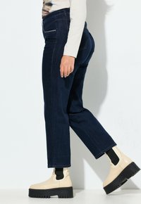 Jeans de pierna recta en azul oscuro hechos de mezclilla, con costuras en contraste y bolsillos laterales, combinados con botas de tobillo beige y calcetines negros.