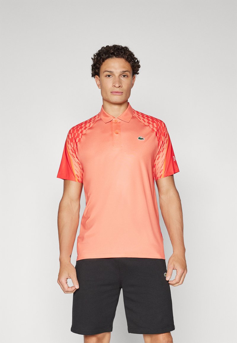 Herren-Poloshirt in einem Farbverlauf von Koralle zu Rot, mit kurzen Ärmeln, gemusterten Akzenten auf den Schultern und einem kleinen grünen Logo auf der Brust.