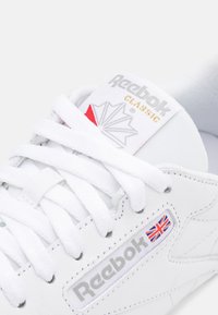 Gros plan d'une basket blanche Reebok Classic montrant les lacets, l'étiquette de la languette avec le logo et le logo latéral avec un petit drapeau Union Jack.