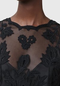 Gros plan sur une personne portant un top noir transparent avec une broderie florale complexe et un décolleté festonné.