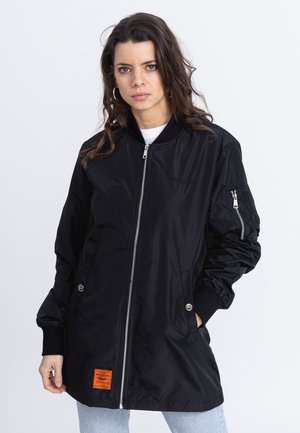Black bomber jacket in nylon with a zip front, side pockets, and ribbed cuffs. Features an orange label at the hem and a smooth texture.