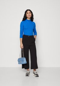 Mingețea albastră cu gât înalt, pantaloni negri largi și adidași cu model. Ținând o geantă din material quiltuit de culoare albastră deschisă, cu un lanț.