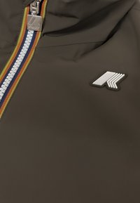 Giacca impermeabile marrone con zip frontale, caratterizzata da una zip bianca con strisce rosse, blu e gialle, e una patch logo argentata.