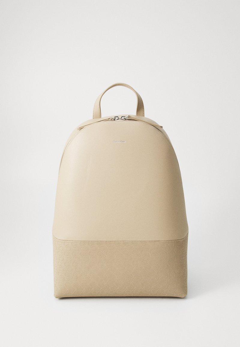 Beige backpack with a smooth upper and textured patterned lower section. Features dual zippers and a top handle for carrying.