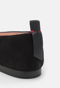 Chaussure en daim noir à enfiler avec un bout arrondi, dotée d'une languette de talon en cuir noir et d'un accent rouge. Surface texturée, design minimaliste.