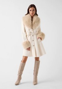 Cream coat with a fuzzy beige collar and cuffs, belted at the waist, paired with knee-high beige boots. Smooth texture, tailored design.