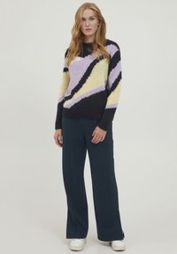 Gebreide trui met marineblauwe, lavendelkleurige en gele zigzagpatronen; ronde hals; geribde boorden; gedragen met donkere wijde broek en witte sneakers.
