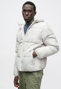 Gai pelēks pūkains jakas ar kapuci, ar rāvējslēdzēja aizdari, ribotām aprocēm un sānu kabatām. Nēsāts virs zilas krekliņas un olīvu krāsas kravjakām.