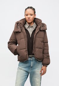 Jonge persoon met gevlochten haar, gekleed in een bruine gewatteerde jas, gestreept shirt, donkere trui en licht versleten spijkerbroek, staand tegen een effen achtergrond.