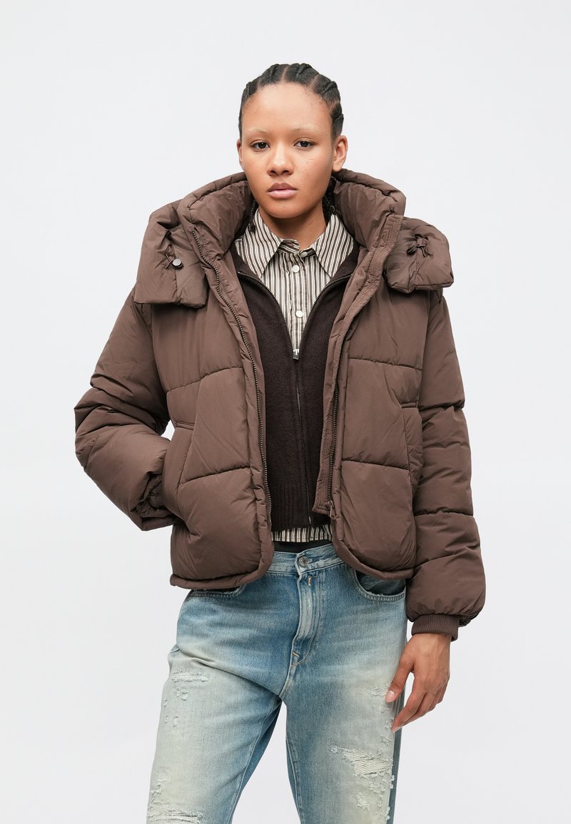 Jonge persoon met gevlochten haar, gekleed in een bruine gewatteerde jas, gestreept shirt, donkere trui en licht versleten spijkerbroek, staand tegen een effen achtergrond.