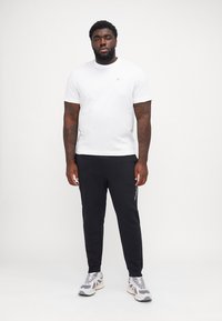 Weißes T-Shirt mit kurzen Ärmeln, schwarze Jogginghose mit seitlichem Streifen, graue Sportsneaker. Das Modell steht mit entspannten Armen an den Seiten vor einem neutralen Hintergrund.