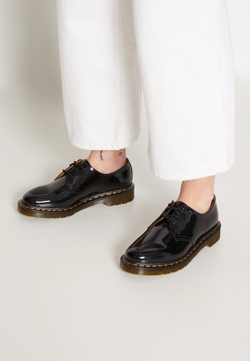 Chaussures à lacets en cuir noir brillant avec des coutures jaunes, portées avec un pantalon blanc court sur un fond clair.