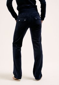 Mörkblå velourbyxor med en figursydd midja, utsvängda ben och två bakfickor med silvriga knapplås. Mjuk textur överlag.