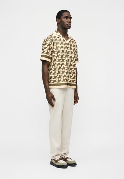 Άνδρας που φοράει μπεζ πουκάμισο με κοντά μανίκια και σχέδιο, ανοιχτό μπεζ παντελόνι και δίχρωμα loafers, στέκεται μπροστά σε απλό ανοιχτό γκρι φόντο.