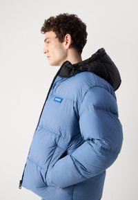 Puffer jacket in light blue with a black hood. Features a zip closure, logo patch, and elastic cuffs. Smooth texture with quilted design.