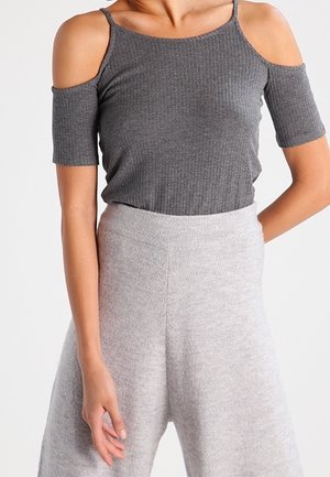 Kvinde iført en grå ribstrikket top med bare skuldre og korte ærmer samt lysegrå, teksturerede bukser med høj talje, stående med den ene hånd bag ryggen.