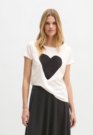 Jonge vrouw met lang blond haar draagt een wit T-shirt met een groot zwart hart en een zwarte rok, tegen een effen achtergrond.