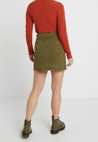 Falda de mezclilla verde oliva con bolsillos traseros, cintura alta y corte recto. Combinada con una camisa roja de mangas largas y botas verdes hasta el tobillo.
