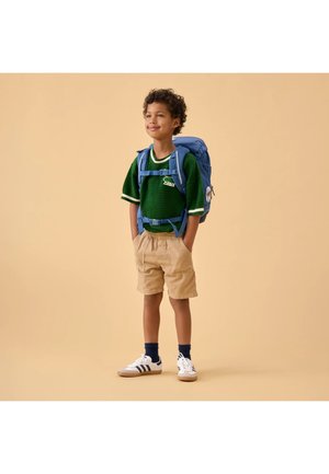 Young boy wearing a green sweater, beige shorts, blue backpack, and white sneakers, standing with hands in pockets against beige background.