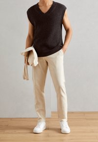 Maglione senza maniche di colore marrone scuro con scollatura a V, abbinato a pantaloni leggeri beige e sneakers bianche, tenuto con una giacca beige.