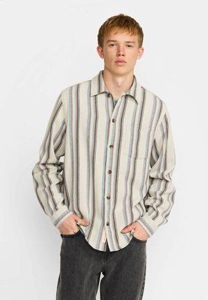 Hombre joven vestido con camisa beige abotonada con rayas verticales azules y naranjas, manos en los bolsillos, de pie frente a un fondo blanco liso.