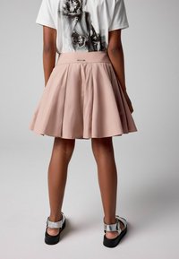 Falda plisada rosa con una textura suave, que presenta una cintura alta y una etiqueta de metal plateado; combinada con sandalias negras y plateadas.
