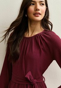 Burgundowa, prążkowana sukienka z długimi rękawami, z zebranym dekoltem i węzłem w talii. Gładka tekstura z subtelnymi plisami.