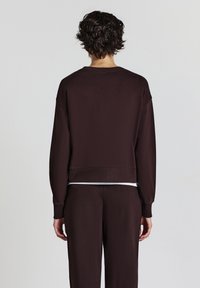 Felpa marrone realizzata in tessuto morbido, con una vestibilità rilassata, maniche larghe e orlo corto. Indossata con pantaloni coordinati.