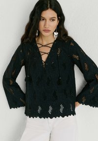 Ženska z dolgimi temnimi lasmi, oblečena v črno čipkasto bluzo z dolgimi rokavi in prepletenim izrezom ter belimi hlačami, stoji ob svetlem zidu.