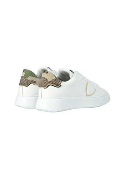 Witte lage sneakers met beige stiksels en groene camouflage-stof op de hiel, weergegeven vanuit een schuin achteraanzicht.