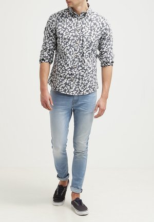 Mężczyzna ubrany w dopasowaną, czarno-białą koszulę w kwiaty na guziki z podwiniętymi rękawami, jasnoniebieskie obcisłe dżinsy i czarne buty sportowe z białymi podeszwami.