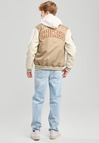 Tan jaqueta varsity com detalhes em creme e a palavra "CHICAGO" em letras laranja. Moleton com capuz branco por baixo, jeans azul claro e tênis brancos.