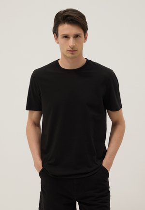 Junger Mann mit dunklem Haar, der ein schlichtes schwarzes kurzärmeliges T-Shirt und schwarze Hose trägt, steht mit den Händen in den Taschen vor einem neutralen Hintergrund.