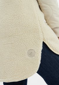 Cremefarbene, strukturierte Fleecejacke mit rundem Logopatch, getragen über dunkelblauer Jeans, mit beigen, gesteppte Ärmeln, die am Arm sichtbar sind.